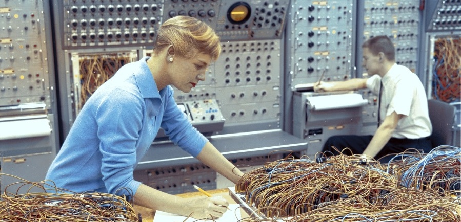 A blond, white woman in a light blue sweater leaning over a fairly chaotic appearing panel of exposed wiring. The image is from 1964.